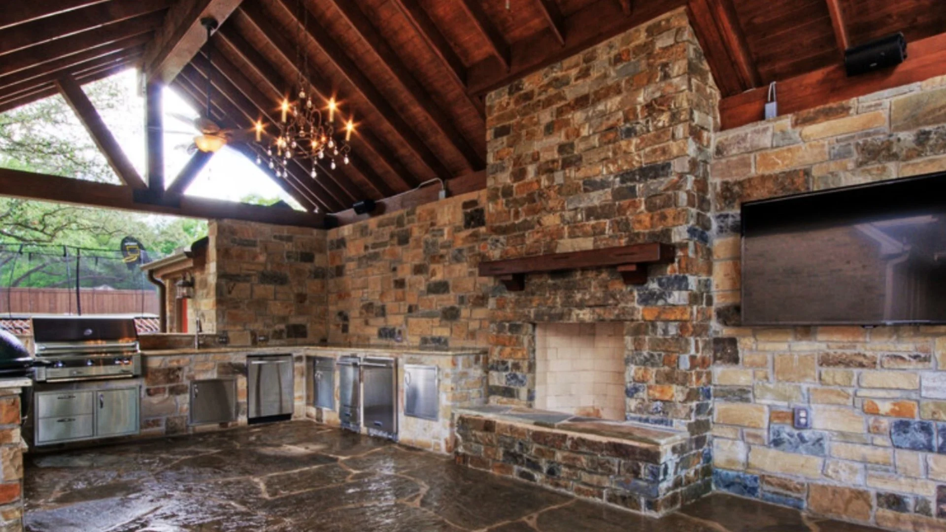 A stunning outdoor kitchen featuring a stone fireplace, equipped with stainless steel appliances, large counters, and beautiful wooden beams overhead, creating a cozy entertaining space.