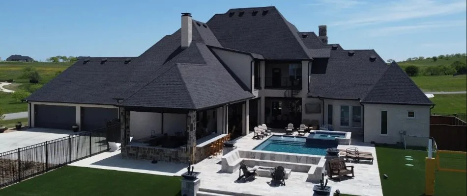 Aerial view of a modern home featuring a spacious patio, swimming pool, and lush green lawn, set against a clear blue sky.