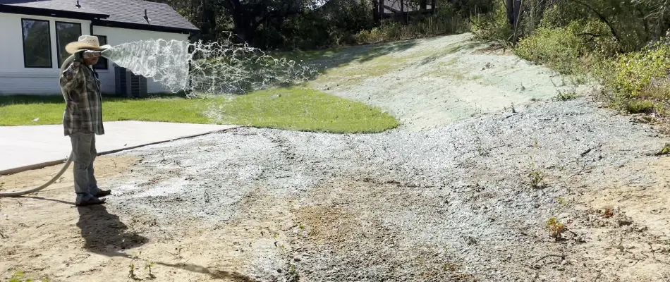 A person in a wide-brimmed hat is seen spraying a mixture of seed, water, and mulch onto a sloped area of soil.