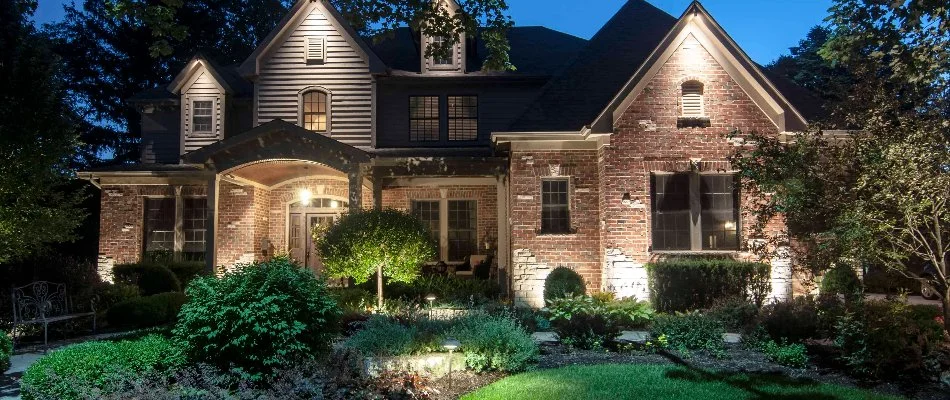 An elegant home beautifully illuminated at night, showcasing meticulous landscaping with lush greenery and decorative plants, under a clear evening sky.