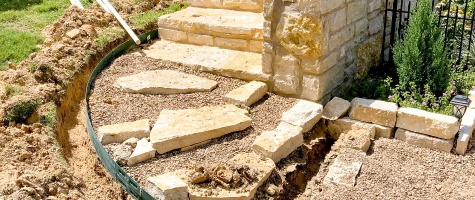 Two landscapers working on a project, installing stone steps and preparing a garden area with decorative rocks and edging, surrounded by freshly disturbed soil and greenery.