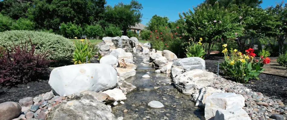 A beautiful landscape featuring a decorative water stream lined with smooth rocks, surrounded by lush plants and colorful flowers under a bright blue sky.