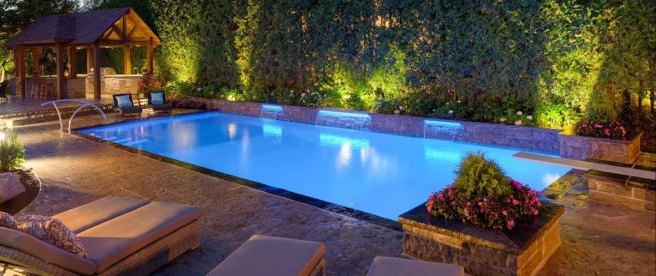  A stunning nighttime view of a luxurious backyard featuring a well-lit swimming pool surrounded by lush greenery.