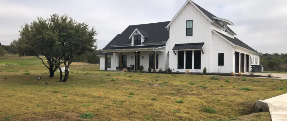 A view of a modern white farmhouse with a black roof, set against a backdrop of rolling hills.