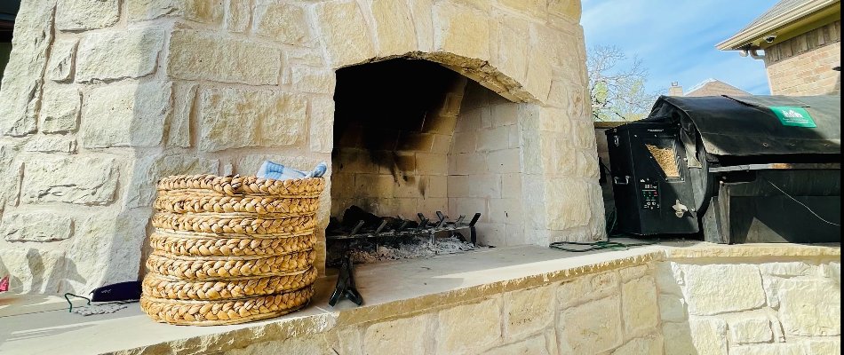 A modern outdoor stone fireplace featuring a wooden mantel and mounted television, enhanced by a sleek design and set against a backdrop of blue skies and nearby grill equipment.