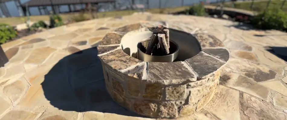 A cozy outdoor fire pit made of stone, featuring logs arranged inside, set on a stone patio overlooking a serene lake in a residential area.