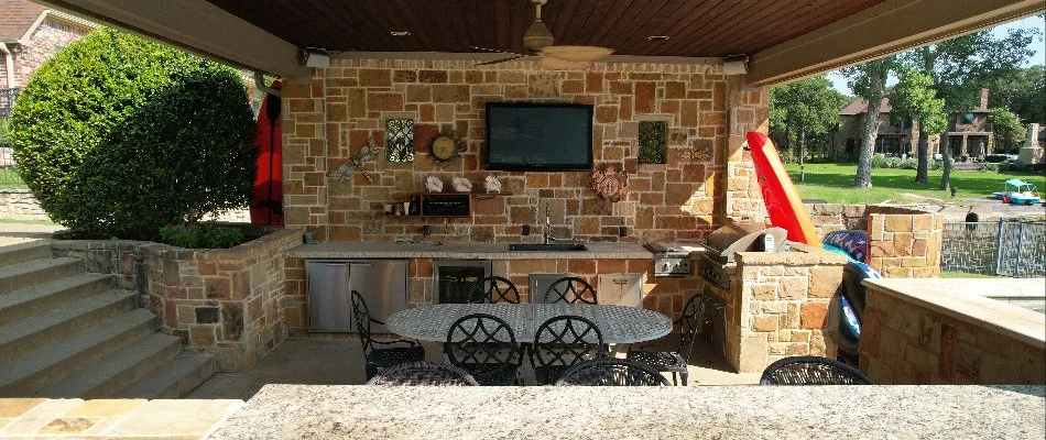 A stylish outdoor kitchen and entertainment area featuring a stone wall, granite countertops, and seating, complete with a grill and television, set in a beautifully landscaped backyard.