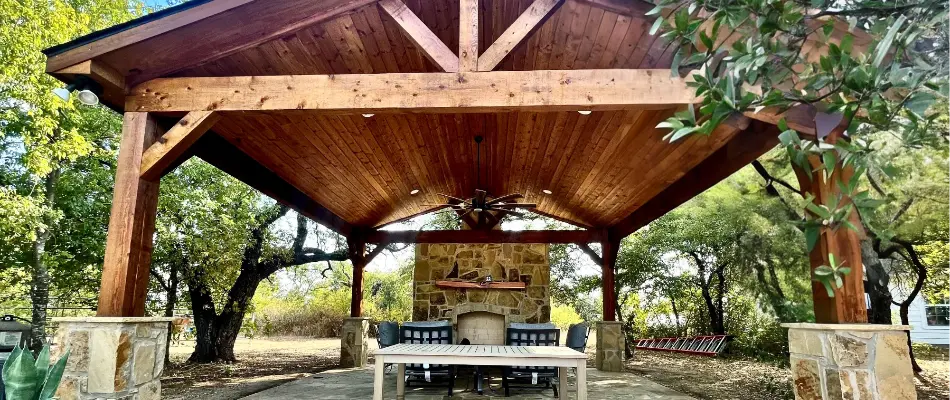 View of an outdoor patio featuring a wooden roof structure and stone columns, with seating and a fireplace area surrounded by trees and landscaped grounds