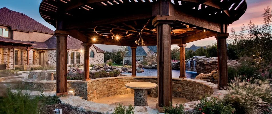 A beautifully designed outdoor pavilion with a stone seating area, surrounded by landscaped gardens and a scenic waterfall, illuminated by soft lights during sunset.