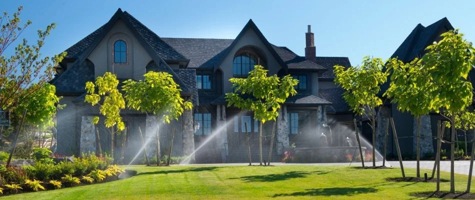 View of a luxurious house with well-maintained landscaping and an irrigation system watering the lawn, featuring green grass and young trees under a clear blue sky.