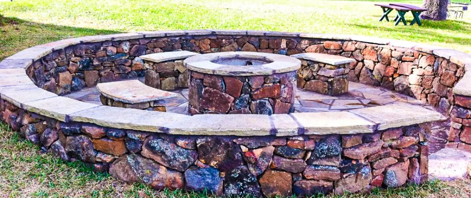 Close-up view of a circular stone fire pit surrounded by seating, set in a grassy backyard area with a wooden structure visible in the background